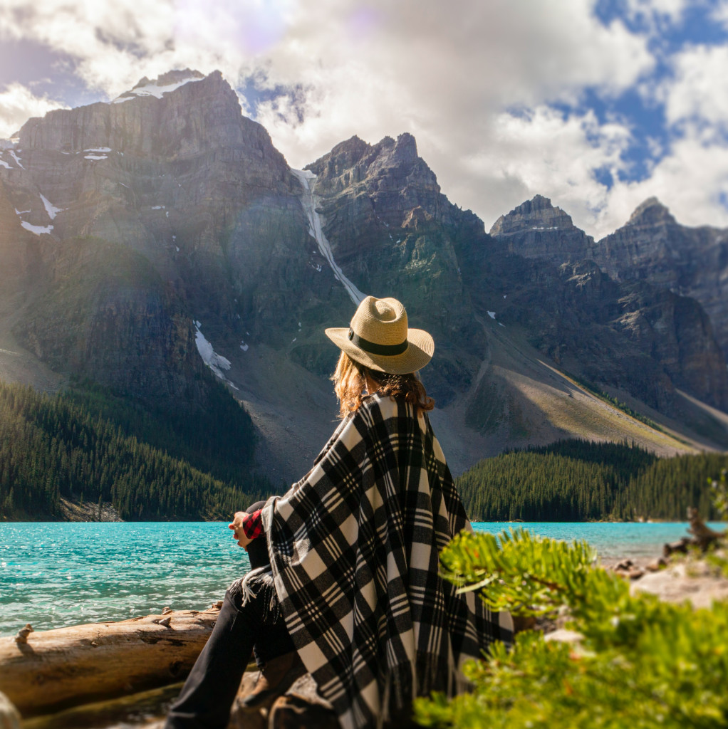 A woman sits on a rock beside a lake looking over at rocky mountains, giving her space to reflect, boost the positives and be encouraged. She is wearing a hat and a checked poncho